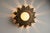 Decorative wall or ceiling light with a radiating sunflower-like metal frame and textured glass diffuser. The fixture is...