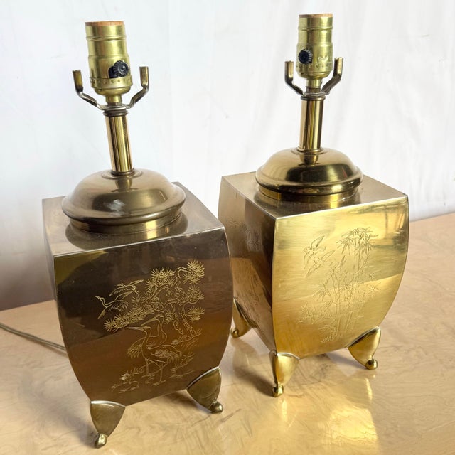 A striking pair of vintage Chinoiserie brass table lamps, richly engraved with serene landscapes, cranes, bamboo, and...