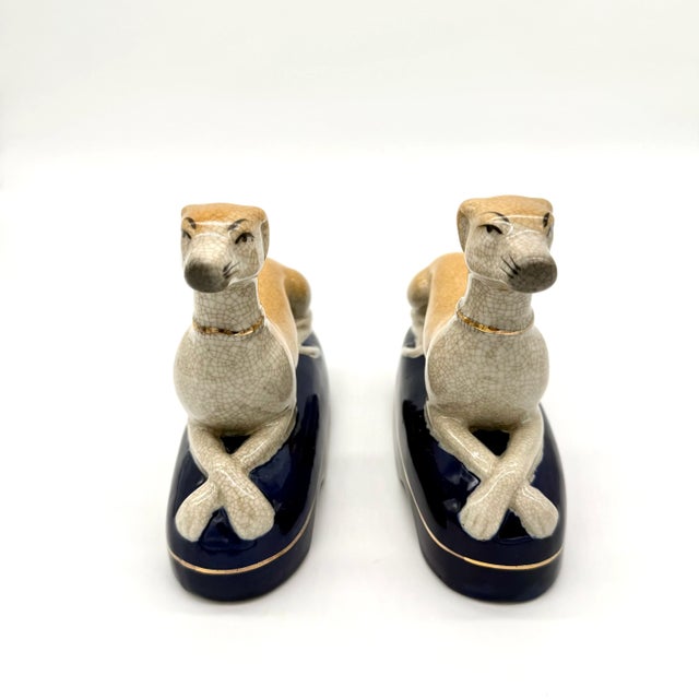 Glazed ceramic reclining greyhounds with crossed paws, featuring a cream crackle glaze, cobalt blue bases, and gilt collar...