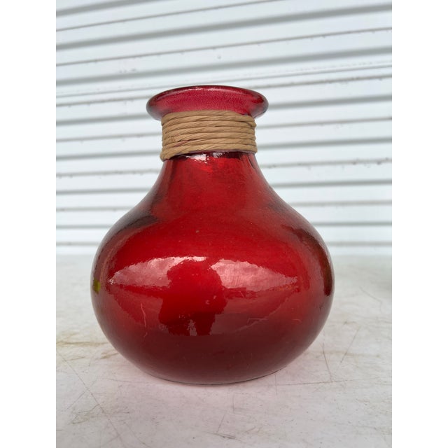 Vintage Red Glass Jug Vase. Has a balloon like shape. Measures 8.5” tall with Rope wrapped around neck. The top rim is 4”...