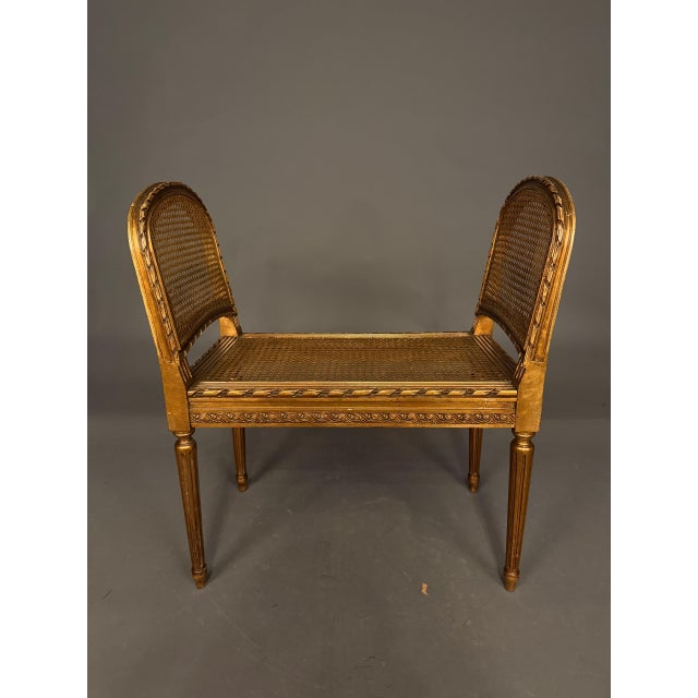A charming 19th-century French Louis XVI-style bench featuring a gilded finish, a hand-carved wooden frame, a cane seat,...