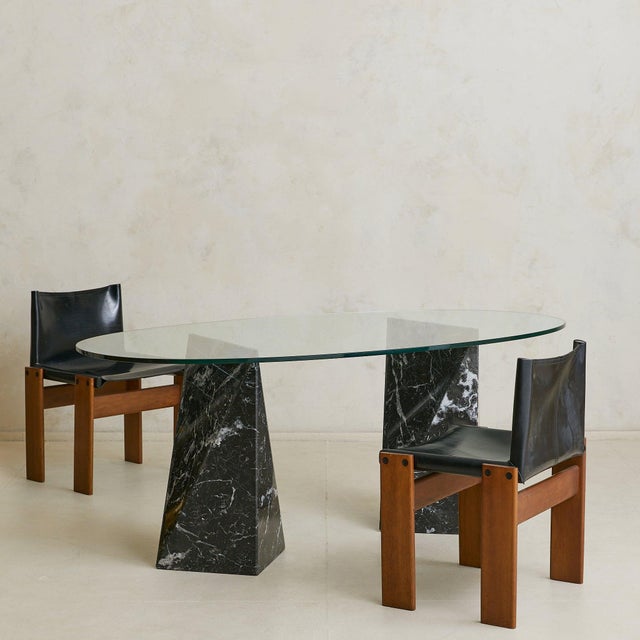An Italian dining table from the 1970s, composed of a thick oval glass top set on two sculptural Nero Marquina marble...
