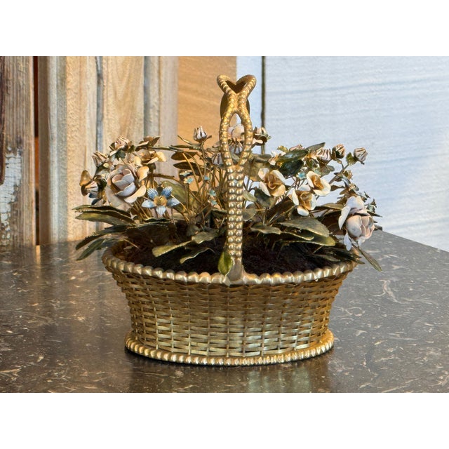 A small decorative basket filled with enameled metal flowers and leaves, arranged within a gold-toned woven basket form...