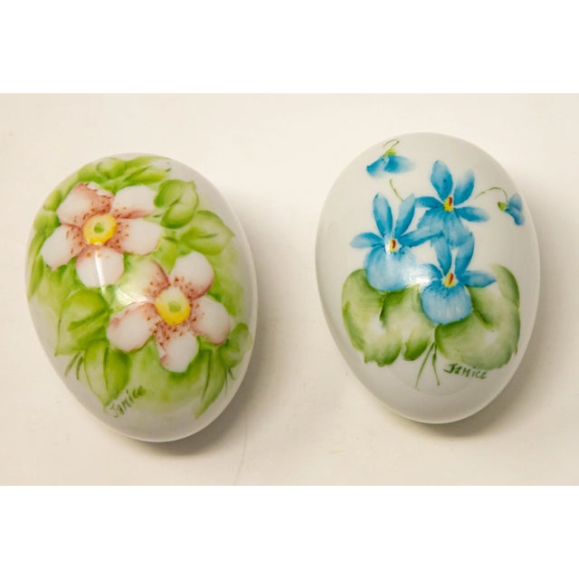 Offering a pair of ceramic hand painted eggs. Both are cream in color. One has light pink florals with greenery around...