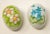 Offering a pair of ceramic hand painted eggs. Both are cream in color. One has light pink florals with greenery around...