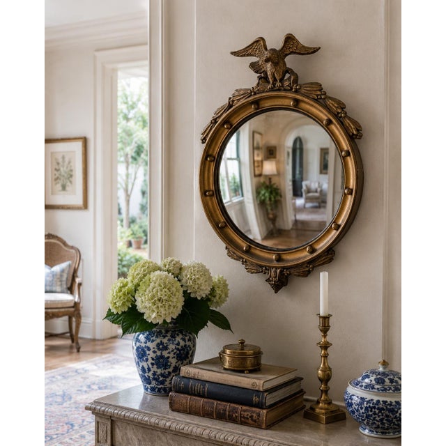 A Federal-style giltwood convex mirror crowned with a carved eagle, its wings outstretched above a laurel motif—an...