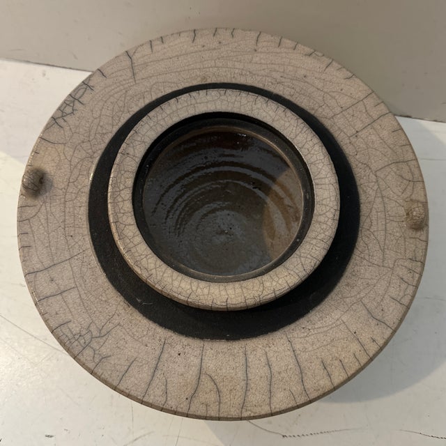 A striking handcrafted raku vessel featuring a softly rounded form and a delicate crackle glaze. Its smoky ivory surface...