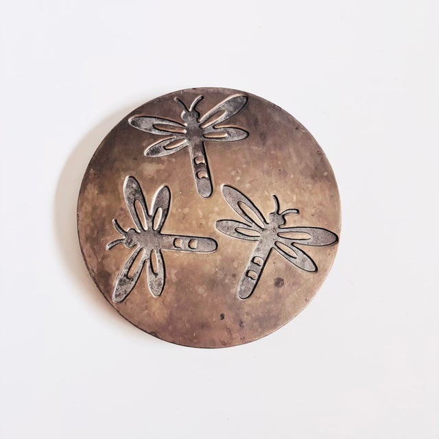 A vintage circular metal trivet. Features an indented dragonfly design and a hoop for hanging as a wall decoration when...