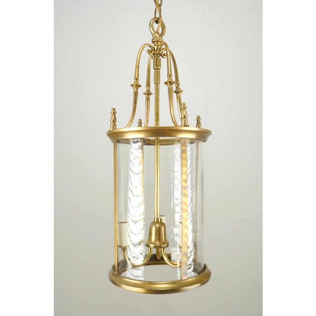 French Neoclassical style ceiling lantern with a brass frame and clear cylindrical glass shade. The chain is made of...