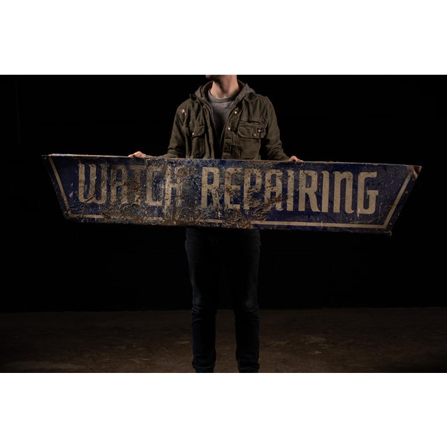 An antique porcelain enamel sign for a watch repair shop. The sign is dark blue with a white font.