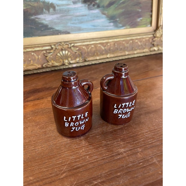 Pair of ceramic salt and pepper shakers shaped as miniature jugs, each with a side handle and hand-painted lettering that...