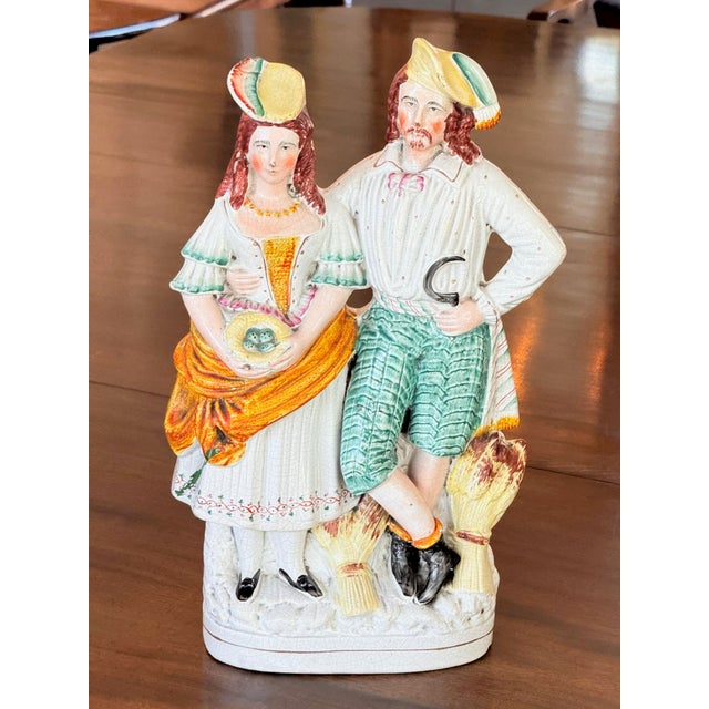 Country couple stand arm in arm on a scrolled base, she with a basket in a yellow bonnet and orange shawl, he with a...