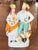 Country couple stand arm in arm on a scrolled base, she with a basket in a yellow bonnet and orange shawl, he with a...