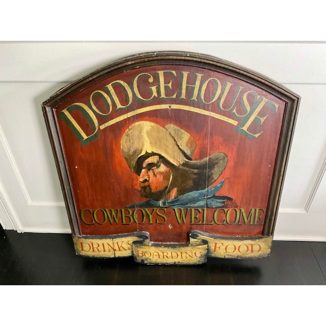 An arch-topped wooden tavern sign painted on a deep red ground, featuring a portrait of a mustached cowboy in profile...