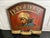An arch-topped wooden tavern sign painted on a deep red ground, featuring a portrait of a mustached cowboy in profile...