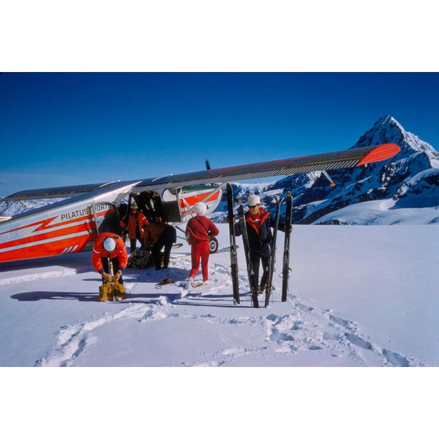 Landing At The Haute Route by Toni Frissell A group of skiers wait by the side of a ski plane at the top of a piste in...