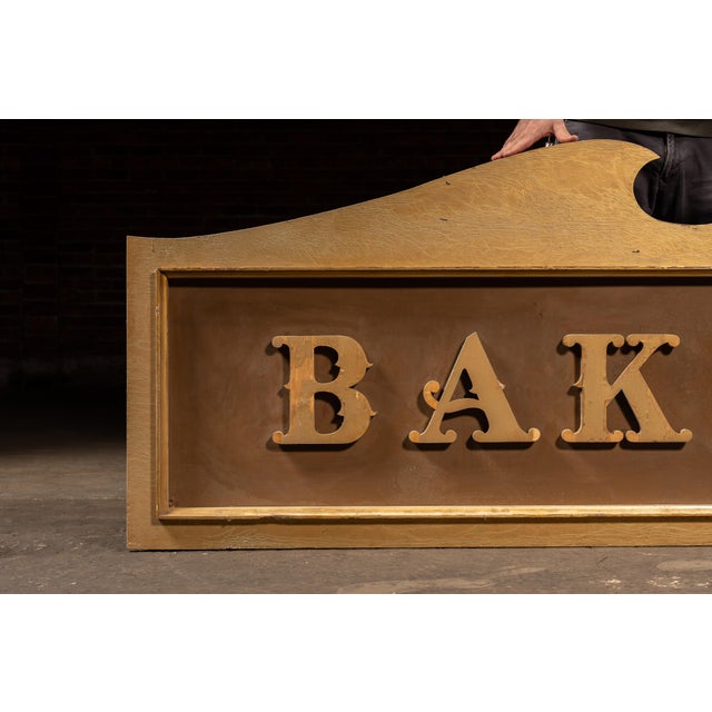 Large hand-crafted wooden storefront sign featuring raised block letters reading BAKERY across a shaped gilt panel. Each...