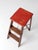 A mid-century industrial folding step ladder featuring a warm wood frame accented by a painted red top—offering a subtle...