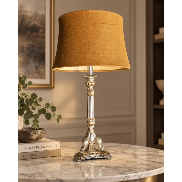 Table lamp with a tall fluted silver-tone column on an ornate base with classical details, paired with a warm brown fabric...