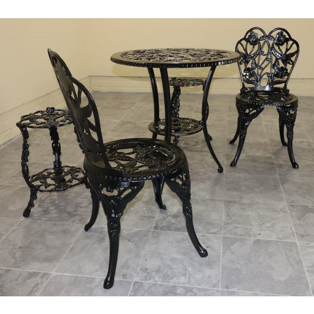 A decorative five-piece garden set featuring two chairs, a round bistro table, and a pair of matching plant stands. Raised...