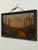 This vintage framed landscape painting depicts a winding path leading through a wooded foreground toward a distant...
