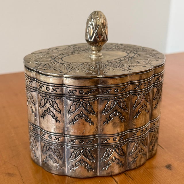 Silverplate lidded trinket box with a pineapple finial and scalloped oval sides. Engraved floral bands and a warm aged...