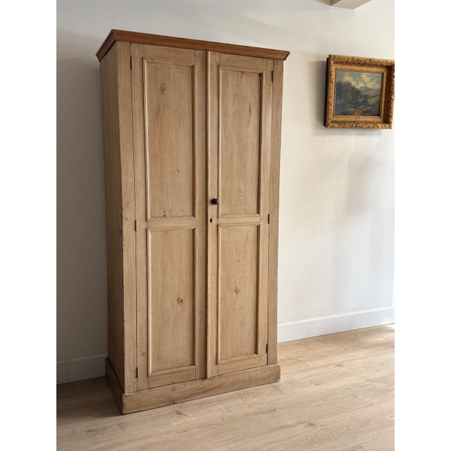 A tall English larder cupboard in pale pine, twin paneled doors with a small turned center knob and a stepped cornice...