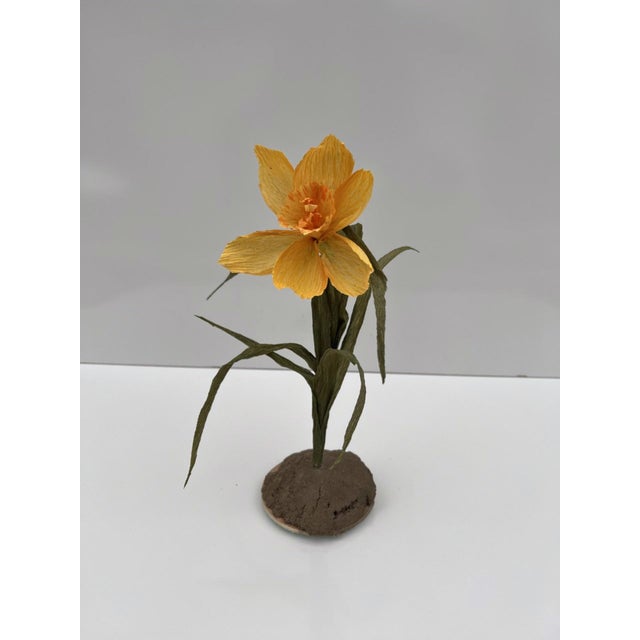 This artwork presents a delicate flower crafted from paper, wire, and paint, emerging from a textured cement base. The...