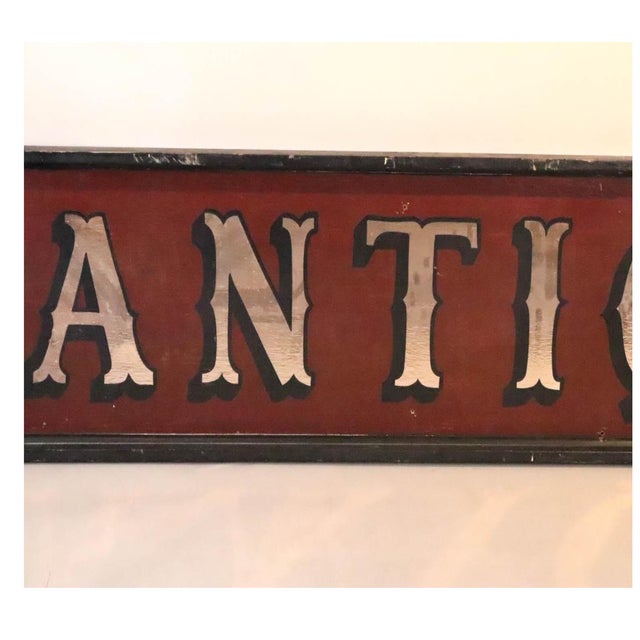 A long, narrow trade sign reading "ANTIQUES" in raised, silver gilded serif letters with worn shading, set against a deep...