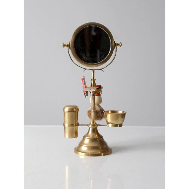 This antique brass grooming stand features a dual-sided mirror with holders for a razor, blades, and brush. The stand...