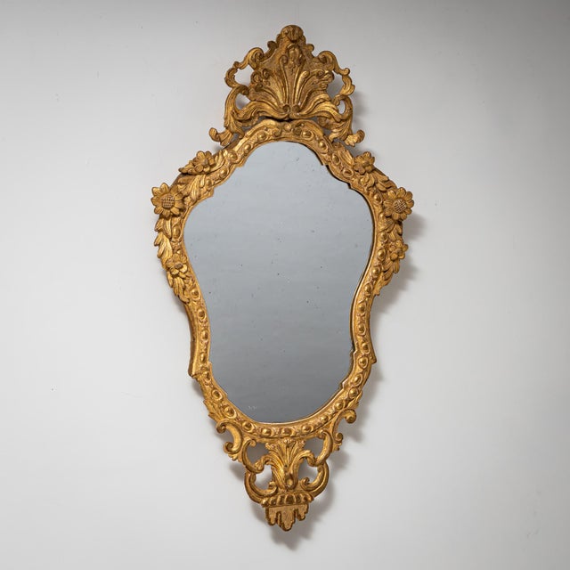 Wall mirror in elongated shield form with a giltwood frame. The frame is richly carved with C-scrolls, shell motifs, and...