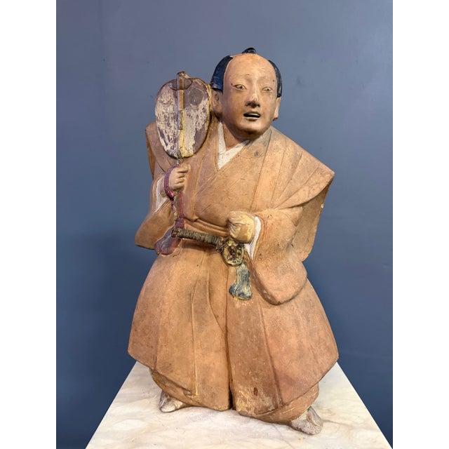 A finely modeled Japanese ceramic figure depicting a samurai or martial attendant holding a gunbai (war fan), dating to...