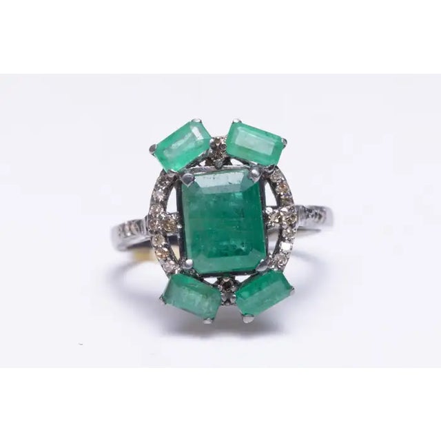 A large center emerald-cut emerald ring flanked with 4 smaller emeralds on the corners. Pave`-set, round faceted diamonds...